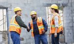 A multi-ethnic group of three workers at a construction site wearing hardhats, reflective vests and safety goggles, meeting and greeting. The Hispanic woman watches as the two men shake hands concept.